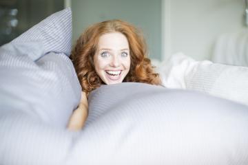 A cheerful red-haired woman peeks out from under striped gray bedding with a wide, joyful smile, creating a playful and bright atmosphere in a cozy bedroom. : Stock Photo or Stock Video Download rcfotostock photos, images and assets rcfotostock | RC Photo Stock.: