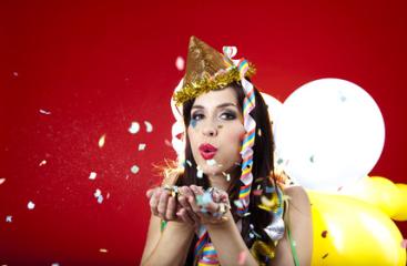 A festive woman wearing a gold party hat and colorful accessories blows confetti with balloons in the background, set against a bright red backdrop, spreading celebration and fun : Stock Photo or Stock Video Download rcfotostock photos, images and assets rcfotostock | RC Photo Stock.: