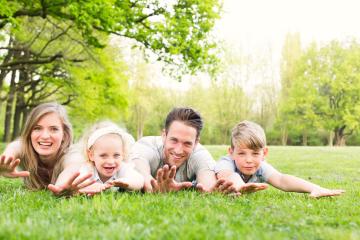 A happy family of four lying on green grass in a sunny park, smiling and reaching forward, surrounded by lush trees and enjoying quality time together in a natural and serene environment- Stock Photo or Stock Video of rcfotostock | RC Photo Stock