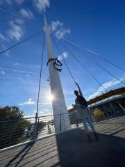 A person in a gray cardigan and jeans stands on a bridge, reaching up towards a colorful horse emblem on a pole, with a bright sun, blue sky, and modern building in the background
- Stock Photo or Stock Video of rcfotostock | RC Photo Stock