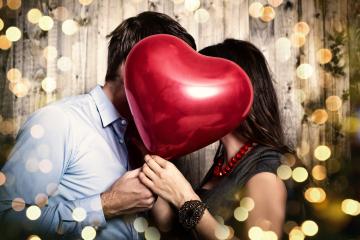 A romantic couple hiding their kiss behind a red heart-shaped balloon, surrounded by glowing bokeh lights and a wooden background, creating a festive and intimate atmosphere : Stock Photo or Stock Video Download rcfotostock photos, images and assets rcfotostock | RC Photo Stock.: