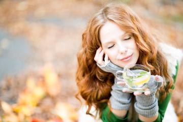 A serene woman with red hair, closed eyes, and a peaceful smile holding a cup of lemon mint tea, sitting outdoors in autumn with colorful leaves in the background, enjoying a cozy moment- Stock Photo or Stock Video of rcfotostock | RC Photo Stock