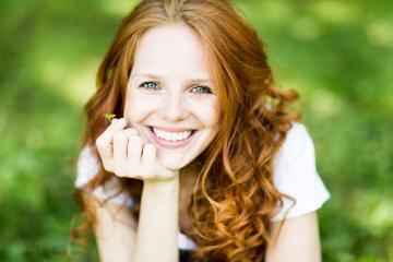 A smiling young woman with red curly hair, bright eyes, and freckles lying on grass, holding a small leaf, radiating happiness, natural beauty, and a connection to nature in a sunny park : Stock Photo or Stock Video Download rcfotostock photos, images and assets rcfotostock | RC Photo Stock.: