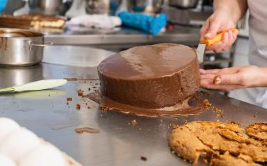 Baker smoothing a chocolate-covered cake with a spatula in a bakery, finishing the cake decoration on a stainless steel work surface in a professional baking environment- Stock Photo or Stock Video of rcfotostock | RC Photo Stock