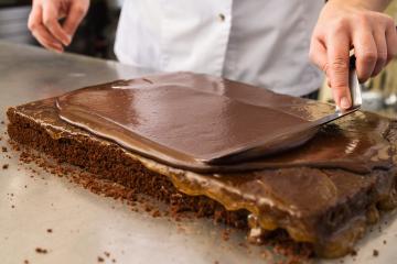 Baker smoothing chocolate glaze on a layered cake with a spatula in a bakery, perfecting the chocolate coating on a caramel-filled cake in a professional baking environment- Stock Photo or Stock Video of rcfotostock | RC Photo Stock
