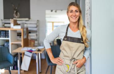 cheerful blonde woman in a gray top and beige apron stands in a tailor workspace. She has a measuring tape and a scissors in her apron pocket and is surrounded by organized craft materials : Stock Photo or Stock Video Download rcfotostock photos, images and assets rcfotostock | RC Photo Stock.: