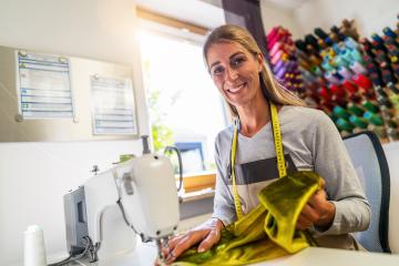 cheerful seamstress woman with a tape measure around her neck is sitting by a sewing machine in a brightly lit workshop, holding a golden silk fabric with colorful spools of thread in the background : Stock Photo or Stock Video Download rcfotostock photos, images and assets rcfotostock | RC Photo Stock.: