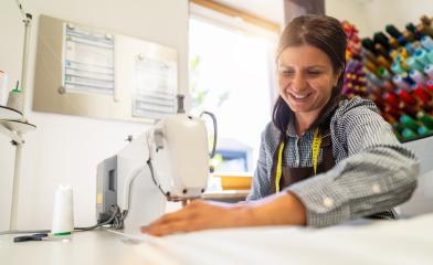 cheerful woman in an apron is working at a sewing machine, stitching white fabric in a well-lit workspace with colorful thread spools in the background- Stock Photo or Stock Video of rcfotostock | RC Photo Stock
