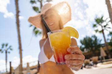 Close-up of a colorful cocktail held by a woman in a bikini, with a straw hat and palm trees at a caribbean island hotel- Stock Photo or Stock Video of rcfotostock | RC Photo Stock