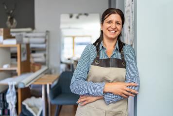 confident woman wearing a checkered shirt and beige apron stands in a workshop with folded arms, smiling towards the camera.- Stock Photo or Stock Video of rcfotostock | RC Photo Stock