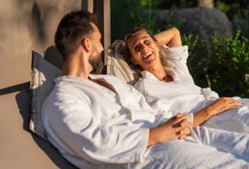 Couple in white bathrobes relaxing and laughing together on lounger in sunlight at a spa wellness hotel- Stock Photo or Stock Video of rcfotostock | RC Photo Stock