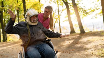 Elderly man in an astronaut helmet extends his arms in a flying gesture, with a nurse smiling beside him in a sunlit autumn park. Dementia retirement home concept image- Stock Photo or Stock Video of rcfotostock | RC Photo Stock