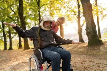 Elderly man in astronaut helmet joyfully extends arms wide, nurse beside him laughing, both in an autumn park. Dementia retirement home concept image- Stock Photo or Stock Video of rcfotostock | RC Photo Stock
