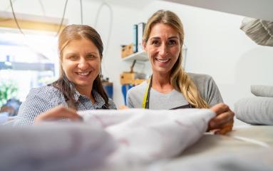 Female work colleagues smiling and closely examining a piece of fabric in a tailoring workshop. One has a measuring tape draped around her neck.- Stock Photo or Stock Video of rcfotostock | RC Photo Stock