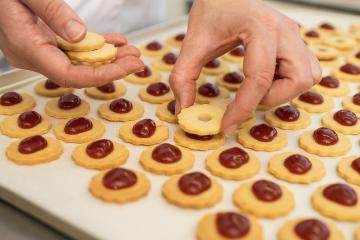 Hands assembling Linzer cookies by placing a top layer with a hole over a bottom cookie with a dollop of red jam. Rows of prepared cookies lie on a baking sheet in the background- Stock Photo or Stock Video of rcfotostock | RC Photo Stock