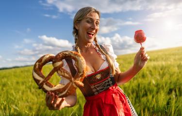Happy Bavarian woman in tracht holding a pretzel and a candy apple, laughing in a sunny wheat field celebrating Oktoberfest ferstival in munich- Stock Photo or Stock Video of rcfotostock | RC Photo Stock