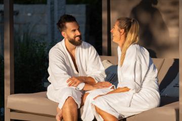 happy couple in white bathrobes sitting and talking on an outdoor bench lounger in sunlight at spa wellness hotel- Stock Photo or Stock Video of rcfotostock | RC Photo Stock