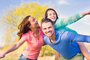 Happy family outdoors enjoying a playful moment, with the father giving a piggyback ride to the daughter while the mother smiles and spreads her arms, set against a sunny, natural background- Stock Photo or Stock Video of rcfotostock | RC Photo Stock