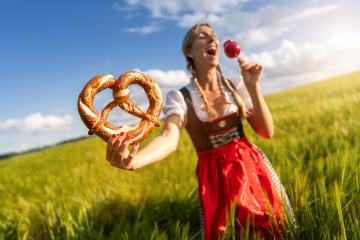 Joyful woman in Bavarian dress holding a pretzel and a candy apple, laughing in a wheat field celebrating Oktoberfest ferstival in munich- Stock Photo or Stock Video of rcfotostock | RC Photo Stock