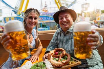 Man and woman in traditional outfits raising beer mugs at a oktoberfest festival, with food on the table and rides in background in munich germany : Stock Photo or Stock Video Download rcfotostock photos, images and assets rcfotostock | RC Photo Stock.: