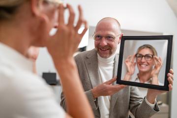 Optician holding a mirror to smiling customer trying on new glasses in optical store.- Stock Photo or Stock Video of rcfotostock | RC Photo Stock