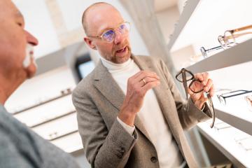 Optometrist presenting eyeglasses to customer in optical shop. He is presenting eyewear selection in front of glasses shelf- Stock Photo or Stock Video of rcfotostock | RC Photo Stock