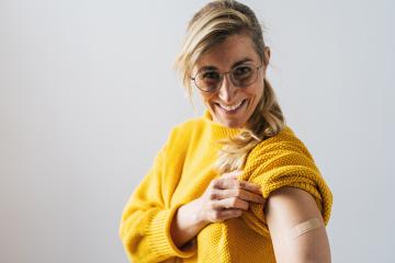 Portrait of a mature woman with glasses smiling after getting a corona vaccine. Woman holding up her shirt sleeve and showing her arm with bandage after receiving vaccination.- Stock Photo or Stock Video of rcfotostock | RC Photo Stock