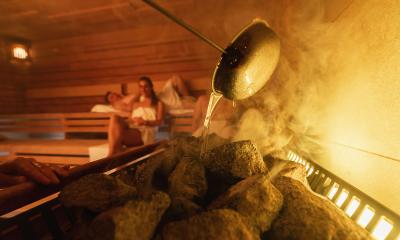 Pouring water on hot stones in a sauna (löyly), steam rising, two people relaxing in the background. Wellness Spa Hotel Conept image.- Stock Photo or Stock Video of rcfotostock | RC Photo Stock