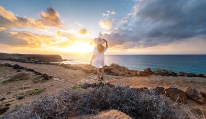 Rear view of a woman in white holding hat looking at the sea during sunset on rocky coast at Playa de Cofete, Fuerteventura, Canary Islands.- Stock Photo or Stock Video of rcfotostock | RC Photo Stock