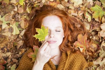 Red-haired woman lying on autumn leaves, holding a maple leaf over one eye, with her eyes closed, embracing the serene and colorful essence of fall in a cozy and peaceful outdoor setting- Stock Photo or Stock Video of rcfotostock | RC Photo Stock