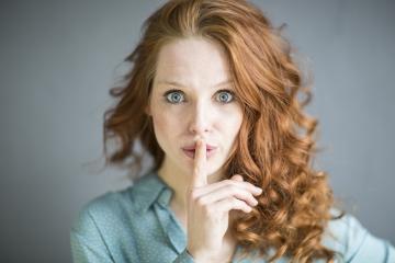 Red-haired woman with striking blue eyes making a quiet gesture by placing her finger on her lips, wearing a green blouse with a subtle pattern, against a soft gray background, expressing secrecy- Stock Photo or Stock Video of rcfotostock | RC Photo Stock