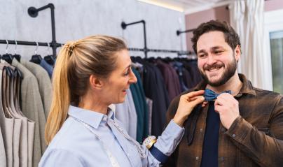 Saleswoman fitting a bow tie on a customer, both are smiling, racks of clothes behind in a men's clothing store : Stock Photo or Stock Video Download rcfotostock photos, images and assets rcfotostock | RC Photo Stock.: