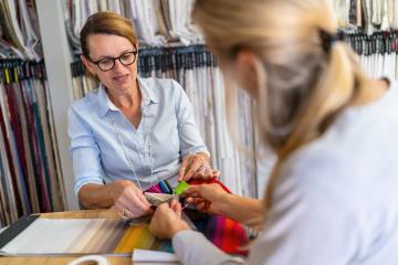 Seamstress sits with her customer in the shop and advises her on choosing her curtain fabric. Seamstress  smiling and touching a color swatch while the other, partly visible, listens attentively.- Stock Photo or Stock Video of rcfotostock | RC Photo Stock