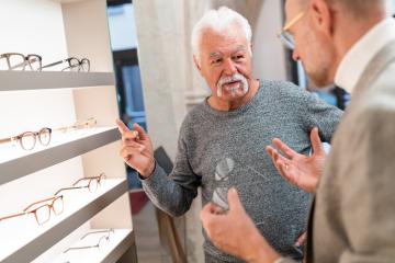 Senior man gesturing during a conversation with an optician in an optical shop with glasses displayed in the background- Stock Photo or Stock Video of rcfotostock | RC Photo Stock