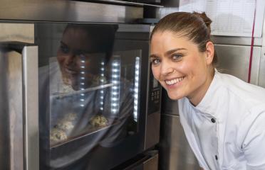 smiling baker in a white uniform is watching cookies bake in the oven. Her reflection is visible on the oven door, adding a touch of anticipation to the baking process. : Stock Photo or Stock Video Download rcfotostock photos, images and assets rcfotostock | RC Photo Stock.: