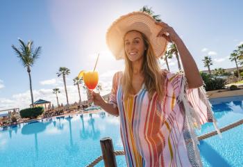 Smiling woman in a straw hat and colorful outfit holding a tropical cocktail by a resort pool with palm trees at caribbean island hotel - Stock Photo or Stock Video of rcfotostock | RC Photo Stock