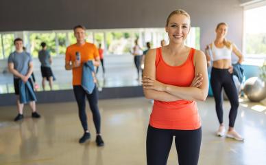 Smiling woman in orange tank top with arms crossed in a gym, people in the background. Teamwork Concept image- Stock Photo or Stock Video of rcfotostock | RC Photo Stock