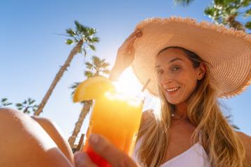 smiling woman wearing a straw hat and holding a tropical cocktail with palm trees in the background at a hotel- Stock Photo or Stock Video of rcfotostock | RC Photo Stock