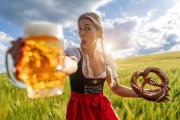 Surprised woman in Bavarian tracht holding beer mug and pretzel in a sunny wheat field ready for Oktoberfest festival in munich, germany- Stock Photo or Stock Video of rcfotostock | RC Photo Stock