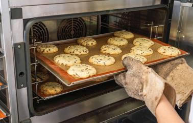 tray of freshly baked chocolate chip cookies is being taken out of the oven using protective oven mitts. The cookies are golden and placed on a silicone baking mat.- Stock Photo or Stock Video of rcfotostock | RC Photo Stock
