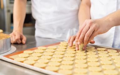 Two bakers in a kitchen are cutting holes in cookie dough shapes on a baking tray. The scene captures the preparation process in a professional baking environment.- Stock Photo or Stock Video of rcfotostock | RC Photo Stock
