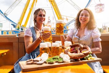 Two women in traditional Bavarian dresses or tracht toasting with beer mugs and  beer and snacks on the table at a festive fairground in Bavaria oktoberfest or dult- Stock Photo or Stock Video of rcfotostock | RC Photo Stock