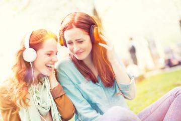 Two young red-haired women wearing headphones share a fun and cheerful moment outdoors, enjoying music together in a bright and sunny park setting.- Stock Photo or Stock Video of rcfotostock | RC Photo Stock