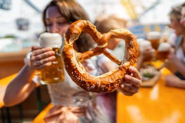 Woman holding a giant pretzel and drinking beer out of a mug with people talking and drinking beer in the background at oktoberfest in munich or  dult in germany : Stock Photo or Stock Video Download rcfotostock photos, images and assets rcfotostock | RC Photo Stock.: