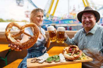 Woman holding a pretzel and toasting beer mugs with a man at a beer tent table with snacks on a wooden board and amusement rides at oktoberfest or dult in germany- Stock Photo or Stock Video of rcfotostock | RC Photo Stock