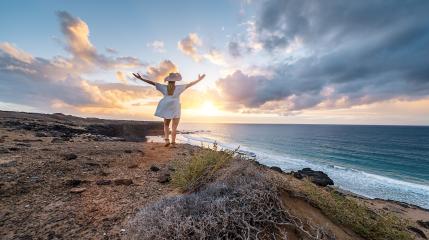 Woman in a white dress and sun hat with arms raised facing the sea on sunset at Playa de Cofete, Fuerteventura, Canary Islands.- Stock Photo or Stock Video of rcfotostock | RC Photo Stock