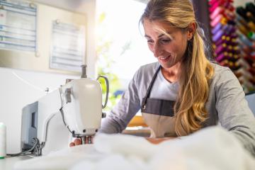 woman in an apron is working at a sewing machine, stitching white fabric in a well-lit workspace with colorful thread spools in the background- Stock Photo or Stock Video of rcfotostock | RC Photo Stock
