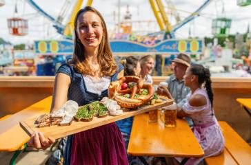woman in Bavarian tracht presenting traditional German snacks (pretzel, obatzter, radishes) at a vibrant fairground dult or oktoberfest in germany- Stock Photo or Stock Video of rcfotostock | RC Photo Stock