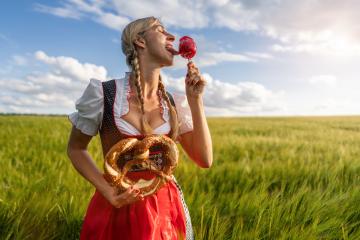 Woman in Bavarian tracht tasting a candy apple and holding a pretzel in a sunny wheat field celebrating Oktoberfest in munich- Stock Photo or Stock Video of rcfotostock | RC Photo Stock