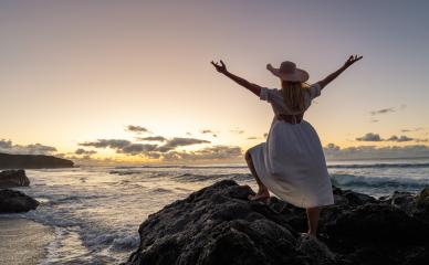 Woman in white dress and straw hat, arms raised, celebrating on rocky beach at sunset with ocean waves- Stock Photo or Stock Video of rcfotostock | RC Photo Stock